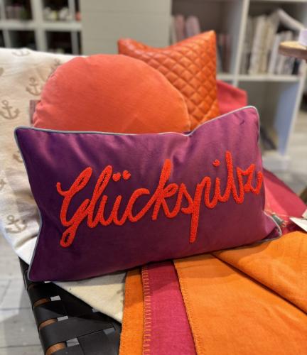 Preview: Kissen Glückspilz von Pad Home in lila mit orangefarbener Stickschrift, dekoriert mit weiteren Kissen in Orange und Pink